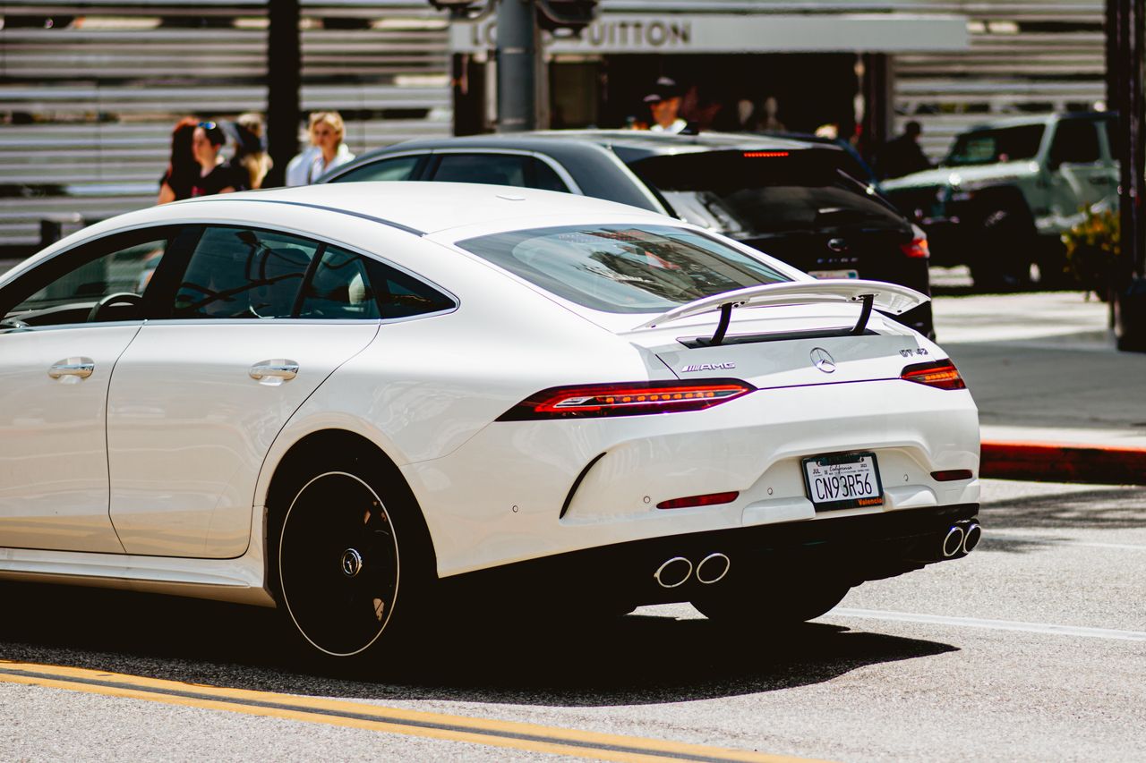A sleek white luxury car cruising in an urban setting with people in the background on a sunny day.